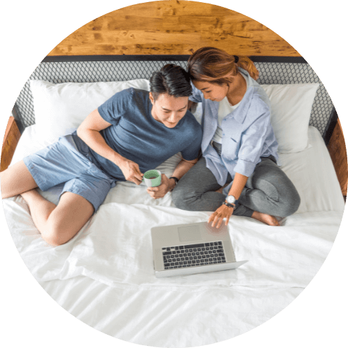 A happy couple sits in bed looking at a laptop together.